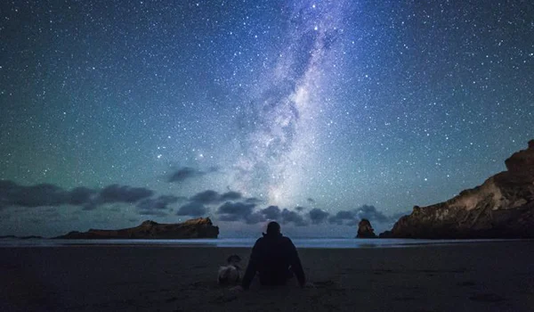 Foto de um homem sentado na praia e com o céu cheio de estrelas e com a Via Lactea.