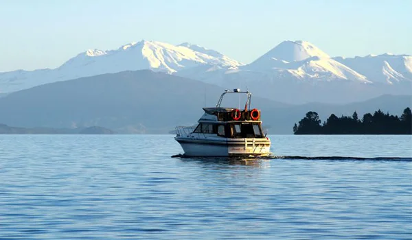 Foto de Lago de Taipo na Nova Zelândia com um barco na água, rodeado de montanhas, algumas nevadas.