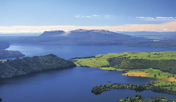 Foto aérea da cidade de Rotorua onde aparece montanhas e terra com vegetação, em contraste com o azul do céu.