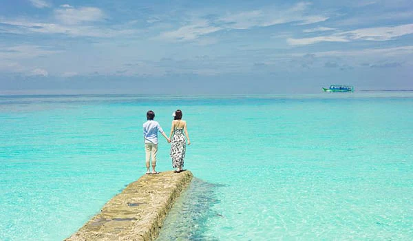 Casal caminhando de mãos dadas em uma ponte de pedra sobre águas cristalinas, com céu azul e uma embarcação ao fundo, criando uma cena romântica e serena em uma praia paradisíaca. Experiência de Casar em Cancun.