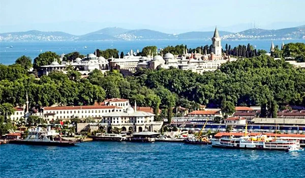 Vista panorâmica de Istambul, na Turquia, com edifícios históricos cercados por vegetação, um minarete ao fundo, barcos atracados no porto e águas azuis em primeiro plano. Ao longe, observam-se colinas e a costa do outro lado do estreito.