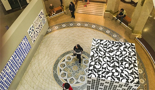 Imagem do lobby de um museu com um chão decorativo, exibição de obras de arte e visitantes apreciando a exposição, destaque para artes visuais variadas.