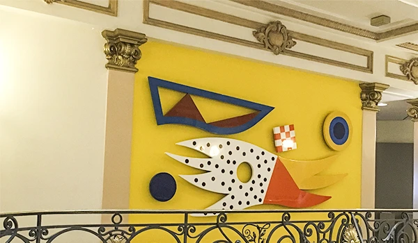 Decoração moderna com elementos artísticos em uma parede amarela, incluindo formas geométricas coloridas e detalhes decorativos clássicos na moldura do teto.