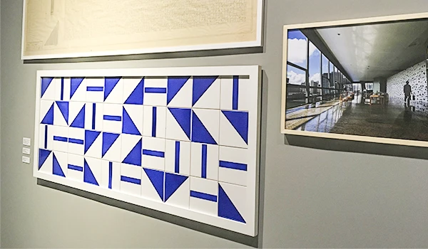 Obra de arte moderna em uma galeria de exposição, composta por formas geométricas azuis e brancas em um quadro grande de moldura branca. À direita, uma imagem de uma exposição com arquitetura contemporânea e uma pessoa vestida de preto caminhando no espaço aberto.