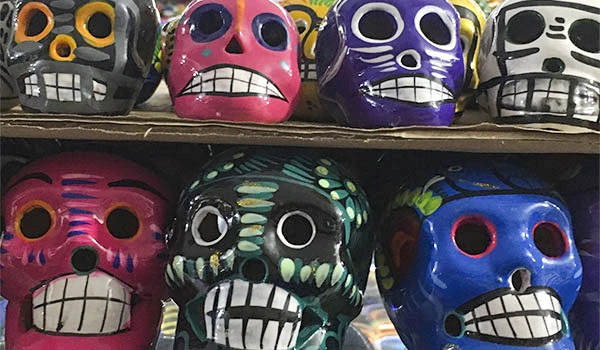 Várias caveiras coloridas de tradição mexicana com expressões variadas, expostas em uma loja ou feira. Decoração típica do Dia de Los Muertos.