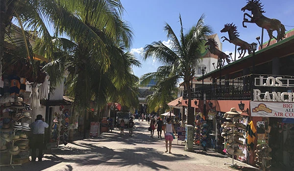 Rua movimentada no centro de Cancun com lojas de souvenirs, caminhantes e palmeiras em um dia ensolarado, próximo ao Los Rancores