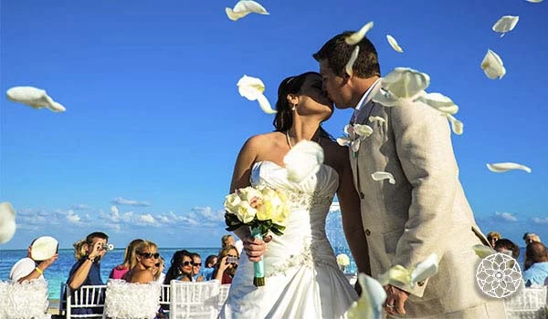 Casal de noivos se beijando em cerimônia de casamento na praia, com convidados jogando pétalas de rosa ao redor e céu azul ao fundo.