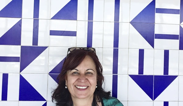 Deusa Rodrigues em frente a um fundo de azulejos azuis e brancos, retratando alegria e estilo brasileiro.