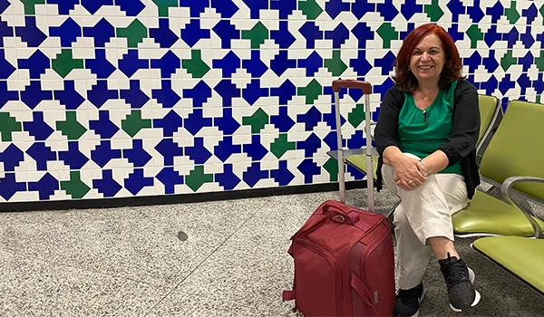 Deusa Rodrigues sentada em um banco de aeroporto ao lado de uma mala vermelha, com um fundo de azulejos coloridos.