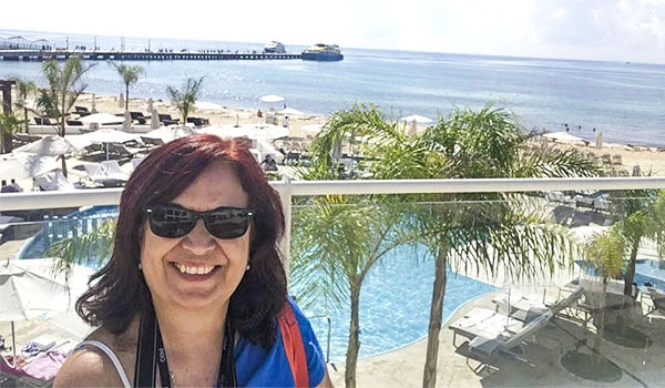 Deusa Rodrigues na piscina do hotel na praia, vestindo óculos escuros e camiseta azul, com vista para o mar e área de praia ao fundo