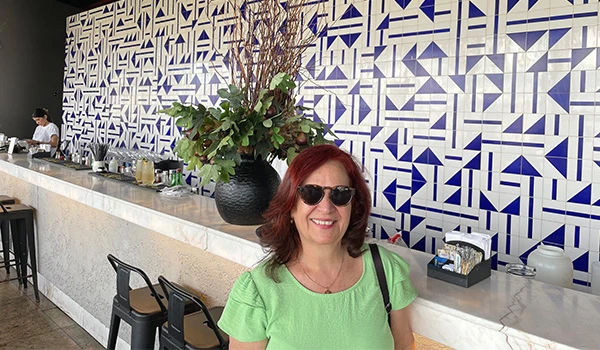 Deusa Rodrigues em um bar moderno com azulejos azuis e brancos ao fundo, decorado com plantas.