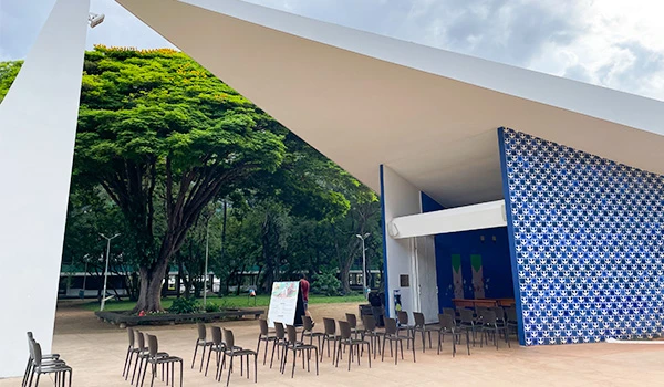 Visão externa de uma moderna estrutura arquitetônica com um telhado em ângulo e parede decorada. O espaço está preparado para um evento, com cadeiras dispostas e um grande árvore ao fundo.