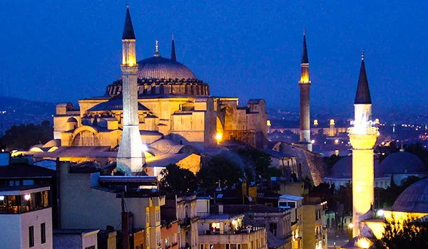 Mesquita de Santa Sofia iluminada à noite, com seus minaretes e cúpulas em destaque contra o céu azul do entardecer, rodeada por edifícios históricos de Istambul, na Turquia.