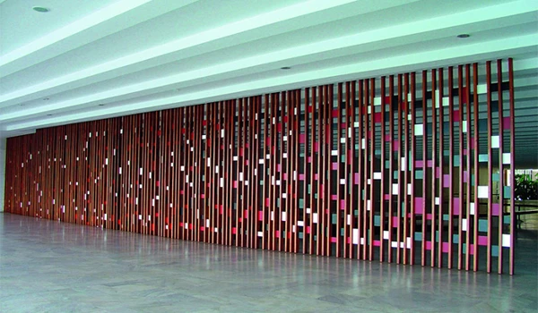 Imagem de um painel decorativo com ripas de madeira verticais em tons de rosa, roxo e branco, instalado em um espaço moderno com parede de vidro ao fundo. Obra de Brasília.