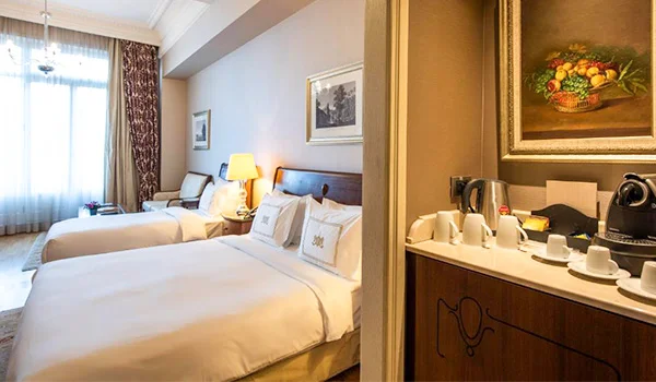 Quarto de hotel elegante com duas camas de casal, roupa de cama branca, cabeceira de madeira, janelas amplas com cortinas e poltronas próximas à janela. À direita, há uma bancada com xícaras, cafeteira e utensílios para preparar bebidas, além de um quadro decorativo de natureza-morta na parede.