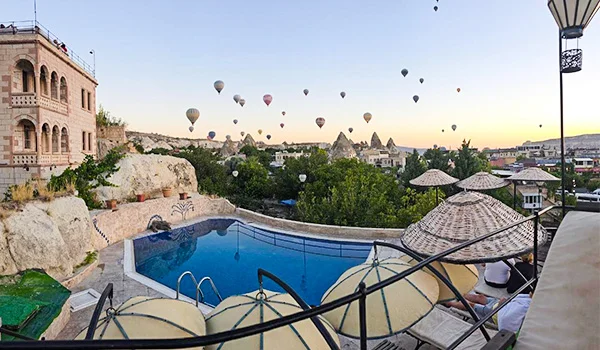 Vista de uma piscina em um hotel na Capadócia, na Turquia, cercada por formações rochosas, construções típicas da região e espreguiçadeiras, com dezenas de balões de ar quente colorindo o céu ao amanhecer.