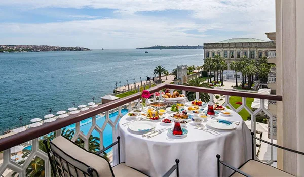Mesa elegantemente posta em uma varanda com vista para o mar, com pratos variados, frutas e taças, em frente a um palácio histórico cercado por palmeiras.