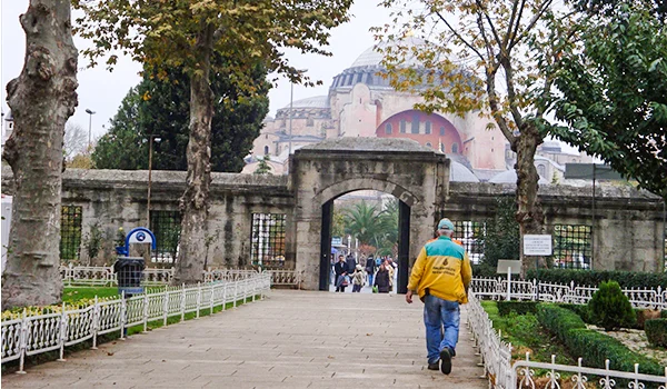 Vista da entrada da Basílica de Santa Sofia, em Istambul, na Turquia, com um portão de pedra, jardins cercados por grades brancas e árvores ao longo do caminho. Ao fundo, destaca-se a arquitetura histórica da basílica, enquanto visitantes caminham em direção ao monumento.