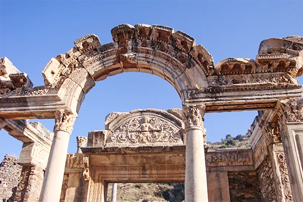 Ruínas antigas de Éfeso, na Turquia, com um arco de pedra ornamentado e colunas romanas diante de um céu azul.