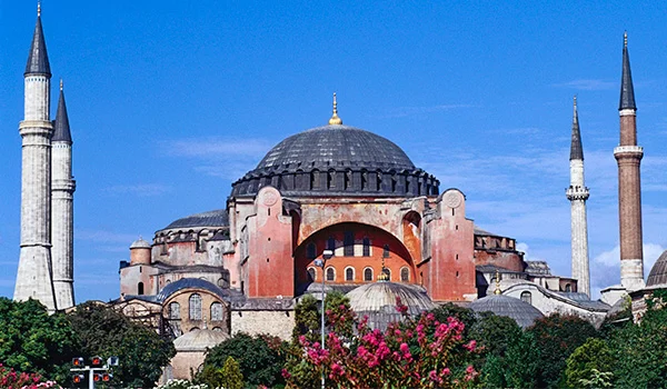 Santa Sofia, em Istambul, na Turquia, com sua grande cúpula central, paredes rosadas, minaretes altos e jardins floridos em primeiro plano, sob um céu azul.