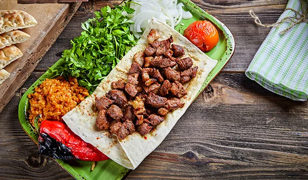 Prato de kebab turco com pedaços de carne grelhada sobre pão achatado, acompanhado de tomate, pimentão assado, arroz temperado, cebola fatiada e salsa fresca, servido em uma travessa verde sobre uma mesa de madeira.
