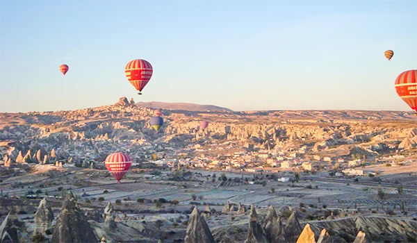 Paisagem da Capadócia, na Turquia, com vários balões de ar quente coloridos sobrevoando formações rochosas, vales e montanhas sob um céu azul-claro.