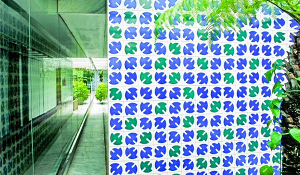 Fachada externa com padrão geométrico em formas de trevos nas cores azul e verde, vista ao lado de um corredor envidraçado com vegetação ao fundo, em ambiente externo.