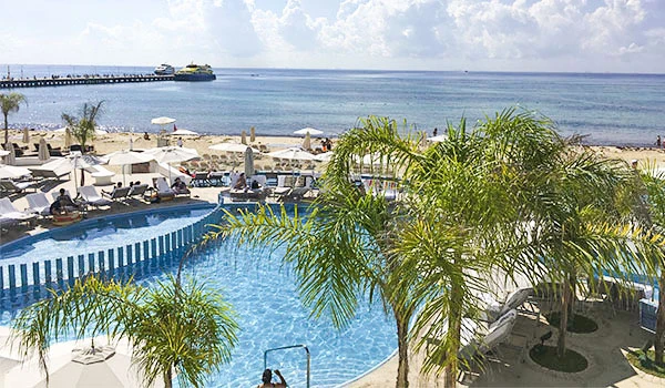 Piscina de resort ao ar livre com vista para o mar, cercada por palmeiras, espreguiçadeiras e guarda-sóis, ideal para relaxamento e lazer.