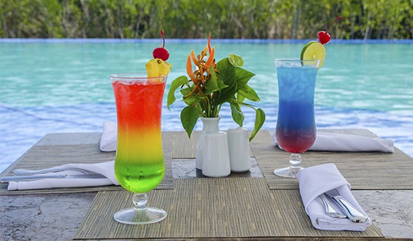 Duas bebidas coloridas com sabores tropicais em copos altos, enfeitadas com cereja, limão e abacaxi, em uma mesa próxima à piscina ao ar livre.