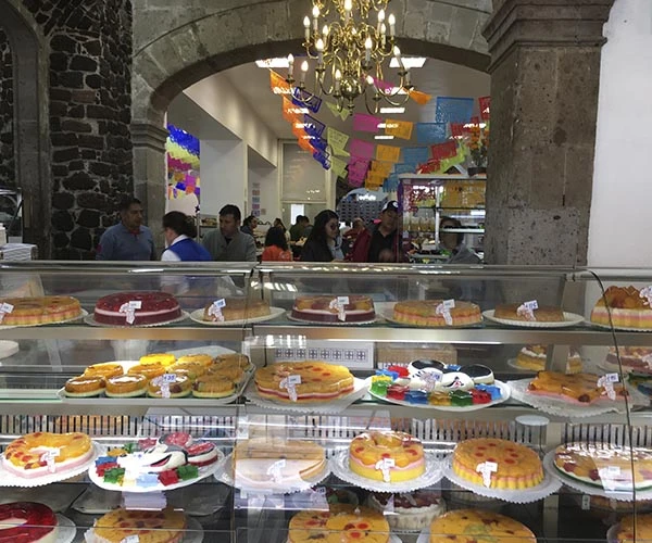 Vitrine de bolos decorados para festa em uma confeitaria no centro histórico, com clientes ao fundo e decoração colorida no teto.