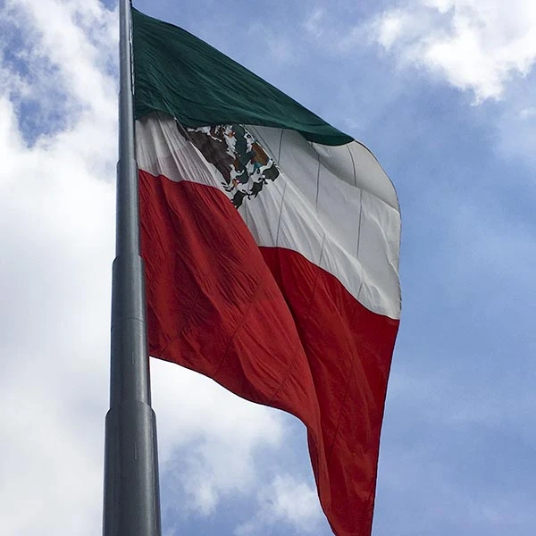 Bandeira do México voando ao vento contra um céu com nuvens, representando patriotismo e orgulho nacional.