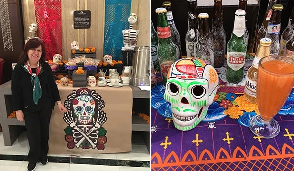 Decoração de Dia de Los Muertos com caveira colorida, bebidas enlatadas e cervejas ao fundo, além de uma mulher sorridente ao lado de uma mesa decorada. Dia dos Mortos no México.