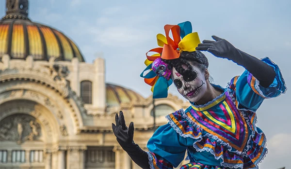 Mulher vestida com traje tradicional de Dia de los Muertos, com maquiagem de caveira mexicana, fantasia colorida com laços, realizando uma apresentação ao ar livre em frente a um prédio histórico. Dia dos Mortos no México.