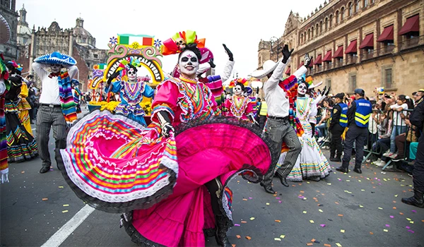 Celebrantes participando do desfile de Dia de los Muertos nas ruas do México, vestindo trajes tradicionais coloridos, com maquiagem de caveira e cenas festivas ao fundo. Dia dos Mortos no México.