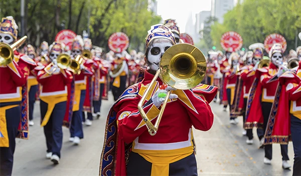 Grupo de músicos em uma marcha carnavalesca, vestidos com fantasias coloridas e máscaras de caveira, tocando instrumentos como trompetes durante Dia dos Mortos no México.
