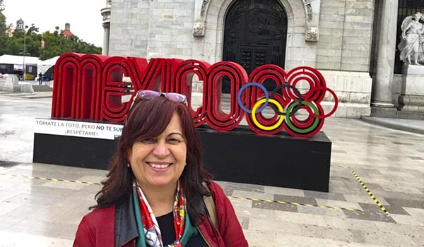 Deusa Rodrigues em frente ao símbolo dos Jogos Olímpicos de Paris 2024 na praça, demonstrando entusiasmo e orgulho. Fundo com monumento histórico ao fundo.