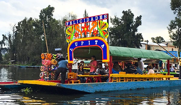 Balsa decorada com tema de felicidades e muitas flores, realizando uma comemoração à beira do rio, com várias pessoas desfrutando.