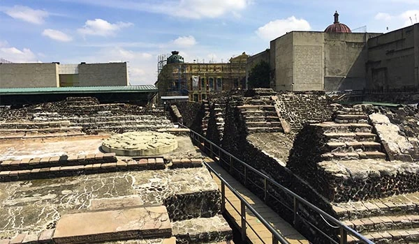 Ruínas de uma antiga estrutura histórica ao ar livre, com degraus de pedra e um fundo com edificações modernas e céu azul. Dia dos Mortos no México.