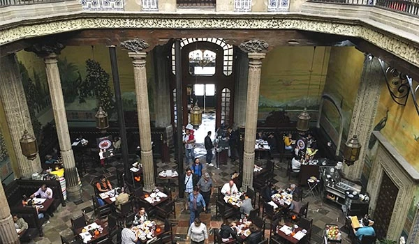 Interior de um restaurante com vários clientes apreciando refeições em mesas, com uma decoração clássica com colunas e janelas grandes, ambiente movimentado.