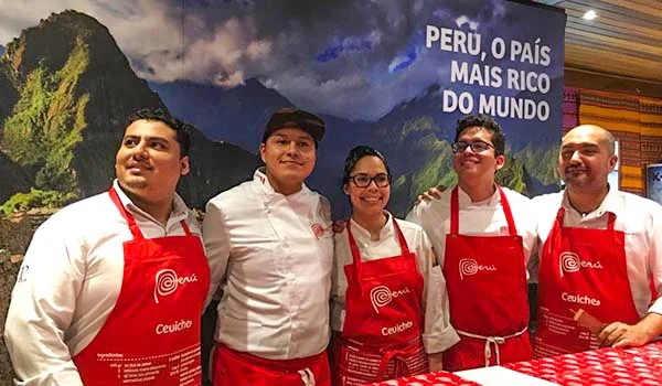 Cinco pessoas posam para uma foto em um evento de culinária peruana, usando aventais vermelhos da marca “Peru”. Ao fundo, há uma imagem de uma paisagem montanhosa do Peru e um texto que diz “PERU, O PAÍS MAIS RICO DO MUNDO”.