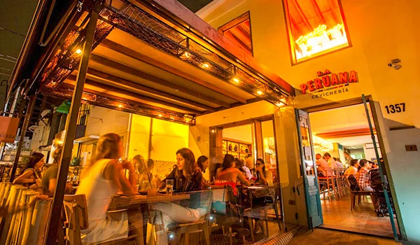 Fachada de um restaurante peruano à noite, iluminada por luzes quentes. Mesas ocupam a área externa e o interior, com vários clientes sentados e conversando. Na entrada, vê-se a placa “La Peruana Cevichería” e o número 1351 na parede.