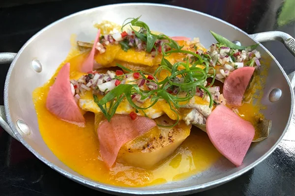 Prato servido em uma pequena frigideira de metal, com peixe e outros ingredientes cobertos por um molho amarelo. A preparação é decorada com cebola roxa picada, ervas verdes e fatias finas de conserva rosada, acompanhada de um pedaço de batata. Restaurante peruano Centro SP.