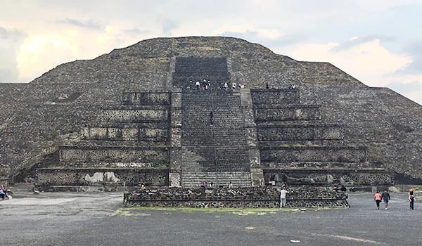 Imagem das Pirâmides do Sol e da Lua, famoso sítio arqueológico Maia, com turistas visitando a estrutura e um céu parcialmente nublado ao fundo.