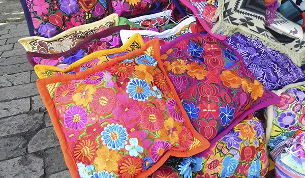 Variedade de bolsas e acessórios femininos artesanais com tecidos coloridos e bordados florais, expostas em uma feira ao ar livre.