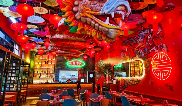 Restaurante de inspiração chinesa e peruana, com decoração predominantemente vermelha, lanternas orientais pendentes, grandes esculturas de dragões e murais decorativos nas paredes. O salão possui mesas e cadeiras, um balcão ao fundo e prateleiras iluminadas com garrafas e objetos decorativos. 