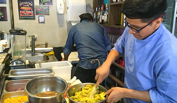 Dois cozinheiros trabalham na cozinha de um restaurante. Em primeiro plano, um homem de óculos e uniforme azul mexe com uma colher em uma tigela com uma preparação amarela. Ao fundo, outro cozinheiro, de avental, trabalha junto à bancada. Sobre as bancadas há recipientes, utensílios e ingredientes, e cartazes coloridos aparecem na parede.
