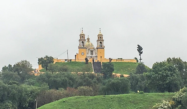 Imagem da igreja histórica no topo de uma colina rodeada por árvores verdes, sob céu nublado em Puebla.