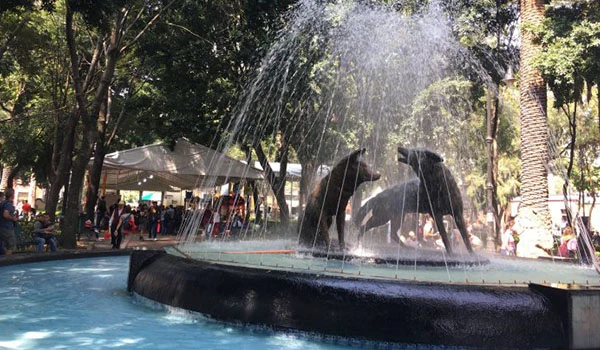 Duas estátuas de golfinhos em uma fonte no parque, com árvores ao redor e pessoas ao fundo, em um dia ensolarado e movimentado.