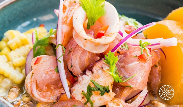 Prato de ceviche com pedaços de peixe e camarões, cebola roxa, milho, tomate, ervas frescas e pedaços de laranja, servido em uma tigela. Restaurante peruano Centro SP.