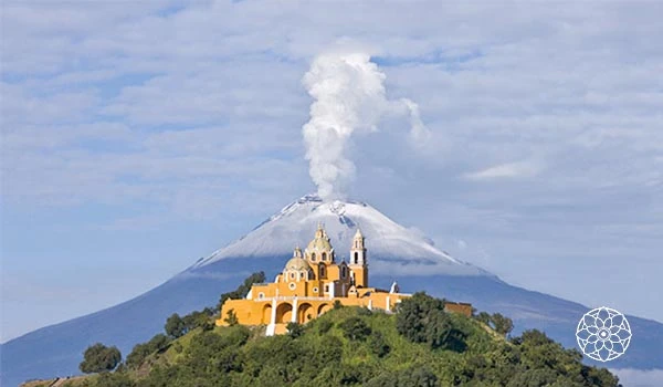 Vista impressionante do vulcão ativo com sua subida de fumaça, ao fundo uma igreja localizada em uma colina verdejante, cenário natural e majestoso.